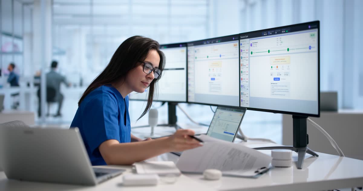 An administrator in scrubs checking paperwork against records on three monitors