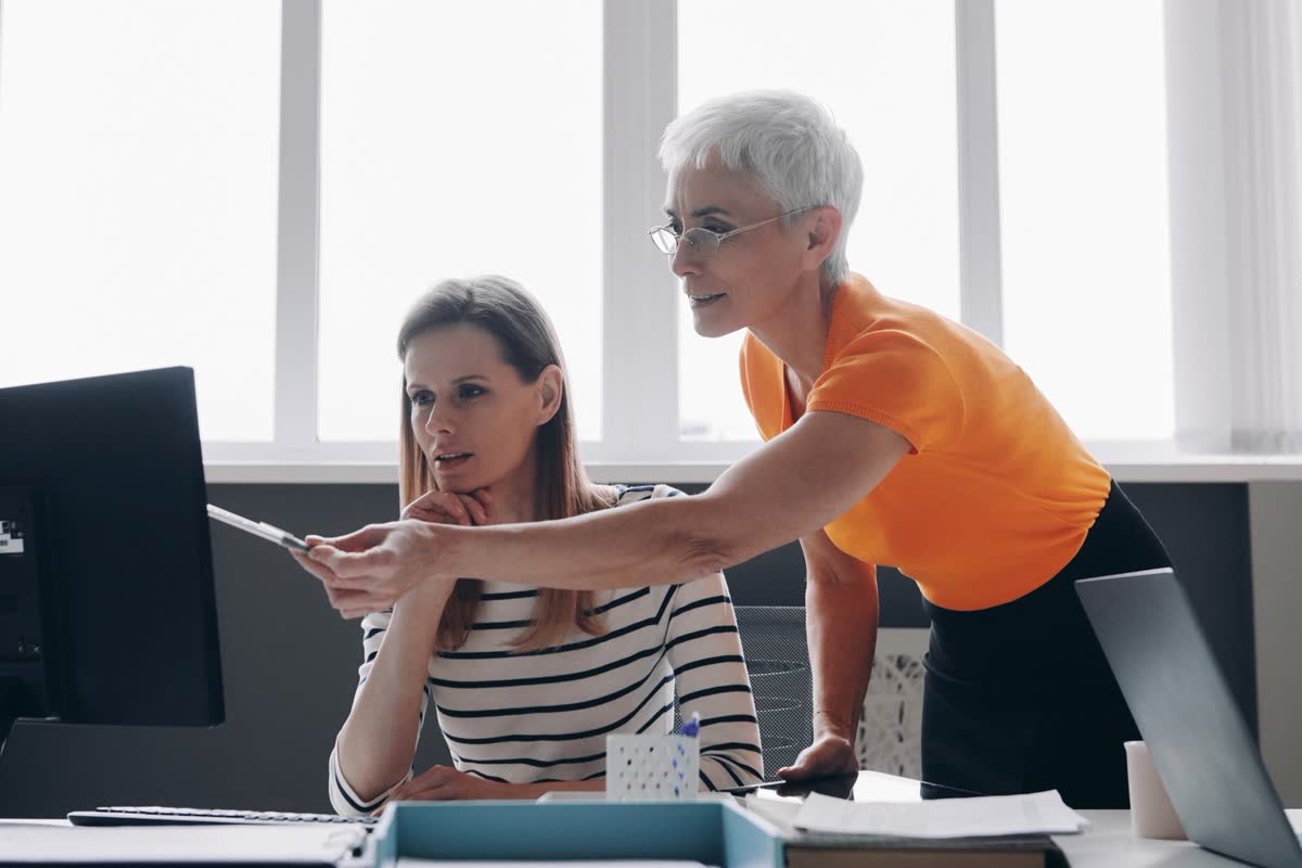 A senior colleague pointing at a monitor while a coworker at the desk follows along