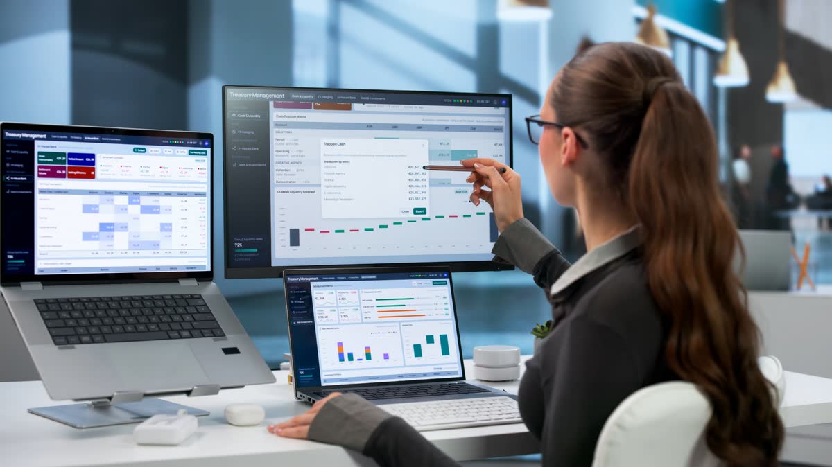 A woman at a desk pointing a pen at a finance dashboard, with a laptop on either side
