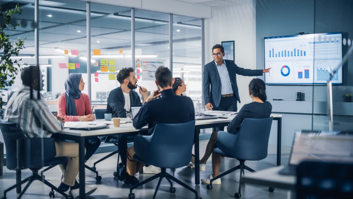 A manager presenting charts on a wall screen to a team around a meeting table