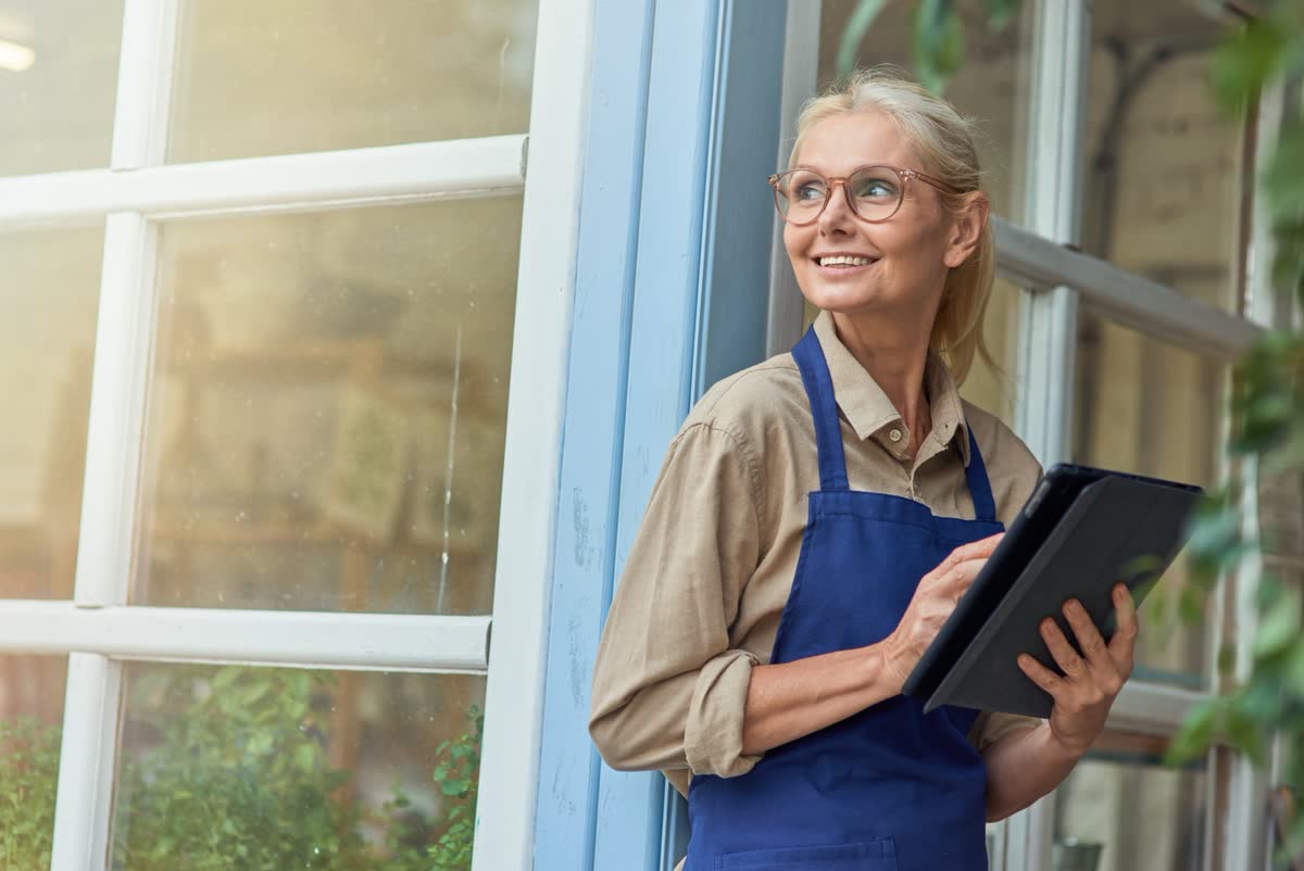 A smiling shop owner in an apron holding a tablet in a sunlit doorway