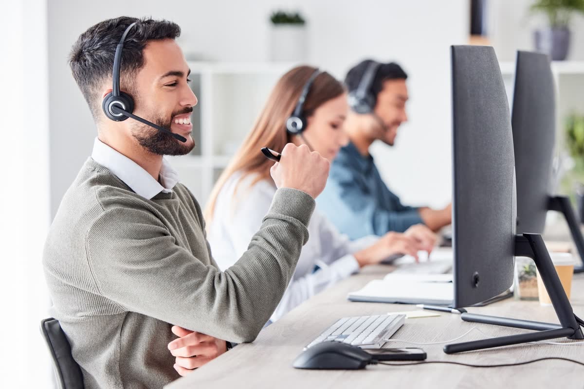 A smiling support agent in a headset at his desk, with colleagues working beside him